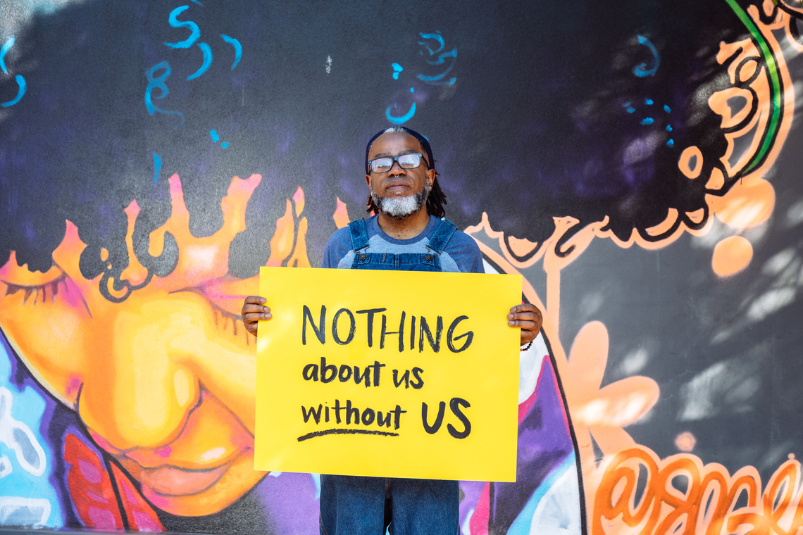 A Deaf Black man wearing glasses looks neutrally at the camera while holding a hand lettered sign declaring &ldquo;NOTHING about us without US.&rdquo;  The man wears denim overalls and has a black and white beard, septum piercing, and hair pulled back with a bandana. The background features a vibrant mural of a Black woman looking down - via Disabled and Here under CC BY 4.0 